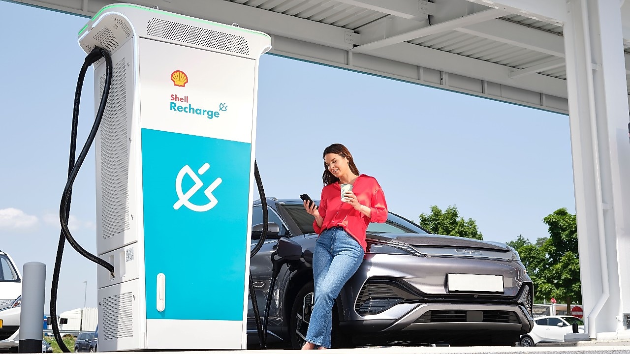 Eine Person benutzt ein Smartphone und lehnt sich an ein schwarzes Elektroauto, das an einer Shell Recharge Ladestation aufgeladen wird