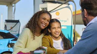 Eine Frau und ein Kind sitzen lächelnd an einem Tisch im Freien an einer Shell Recharge Ladestation. Ein Mann sitzt ihnen gegenüber. Im Hintergrund ist ein Elektrofahrzeug an einer Ladesäule zu sehen.
