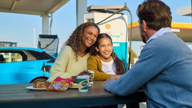 Eine Frau und ein Kind sitzen lächelnd an einem Tisch im Freien an einer Shell Recharge Ladestation. Ein Mann sitzt ihnen gegenüber. Im Hintergrund ist ein Elektrofahrzeug an einer Ladesäule zu sehen.