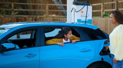 Frau lädt ein blaues Elektroauto an einer Shell Recharge Ladesäule, während ein Kind aus dem geöffneten Autofenster schaut.