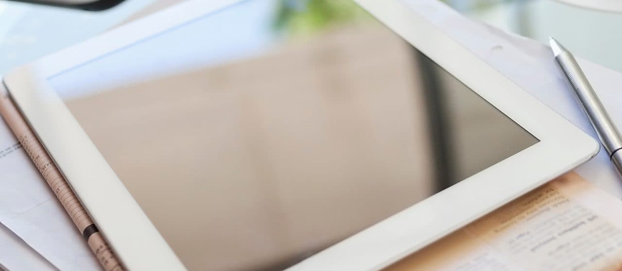 Ein Tablet liegt auf dem Tisch neben einer Tasse Tee, Zeitungen und einem Stift 