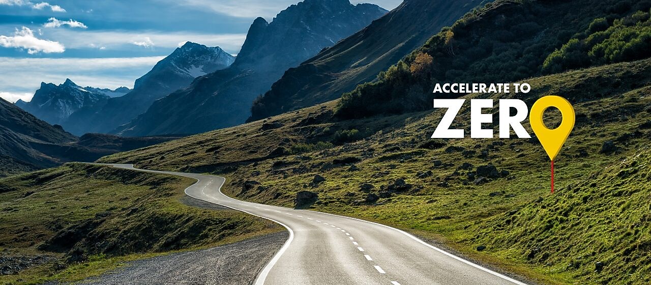 Auto fährt auf einer Schnellstraße durch grüne Landschaft mit Bergen im Hintergrund, Textaufschrift: "Accelerate to Zero".
