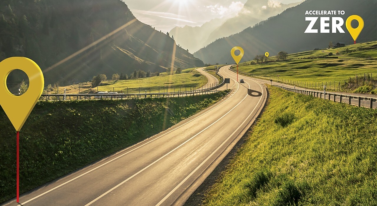 Auto fährt auf einer Schnellstraße durch grüne Landschaft mit Bergen im Hintergrund, Textaufschrift: "Accelerate to Zero".