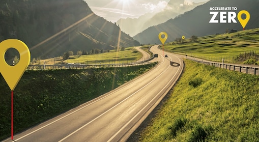 Auto fährt auf einer Schnellstraße durch grüne Landschaft mit Bergen im Hintergrund, Textaufschrift: "Accelerate to Zero".