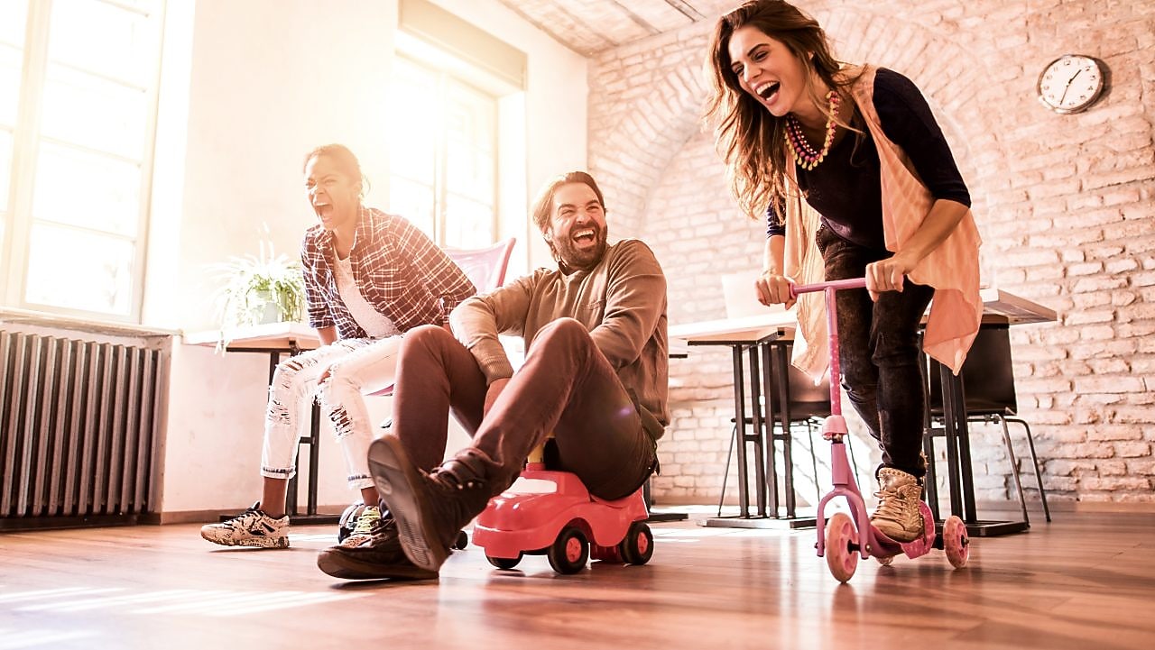 3 Personen fahren mit Spielzeugrollern und Spielzeugautos durch ein Zimmer.