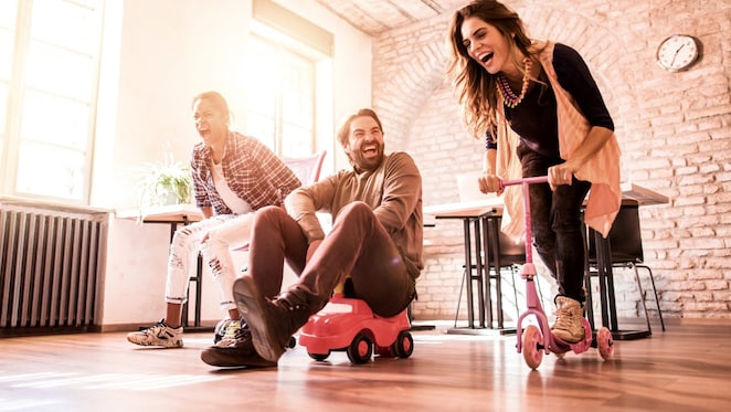 3 Personen fahren mit Spielzeugrollern und Spielzeugautos durch ein Zimmer.