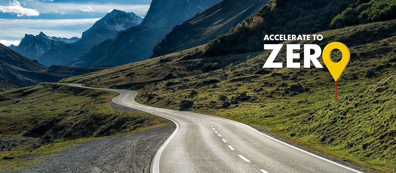 Auto fährt auf einer Schnellstraße durch grüne Landschaft mit Bergen im Hintergrund, Textaufschrift: "Accelerate to Zero".