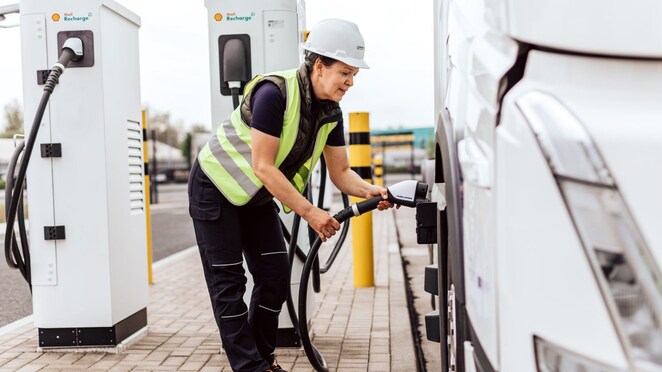 Eine Lkw-Lenkerin mit Warnweste und weißem Helm steht an einem Shell Recharge Depot und steckt gerade den Ladestecker in den E-Lkw.