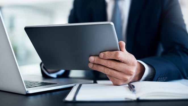 Ein Mann im Anzug sitzt am Schreibtisch und hält ein Tablet in der Hand. Daneben der offene Laptop und ein Notizbuch.