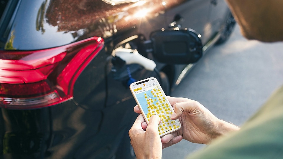 Im Vordergrund nutzt ein Fahrer das Smartphone mit der NewMotion App zur Anzeige der verfügbaren Ladepunkte. Im Hintergrund lädt das E-Fahrzeug an einer E-Mobility Station.
