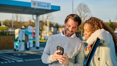 in Mann und eine Frau stehen lächelnd vor einer Shell Recharge Ladestation und schauen gemeinsam auf ein Smartphone. Im Hintergrund sind mehrere türkisfarbene Ladesäulen zu sehen.