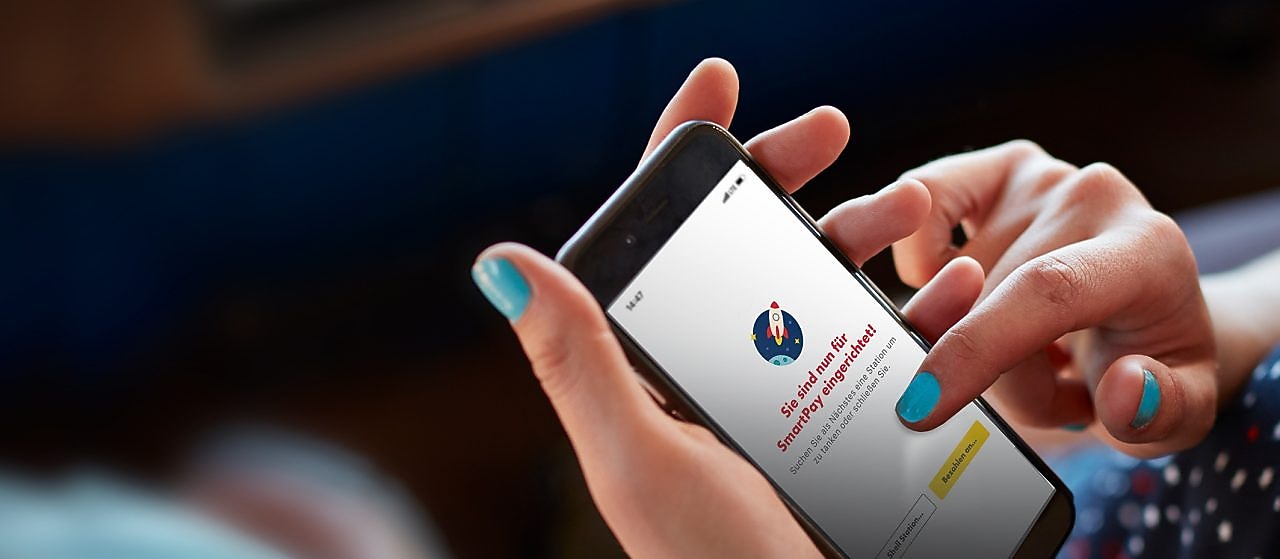 Frau sitzt im Auto und hält in der Hand ein Handy mit geöffneter Shell App und fertig eingerichtetem Shell SmartPay