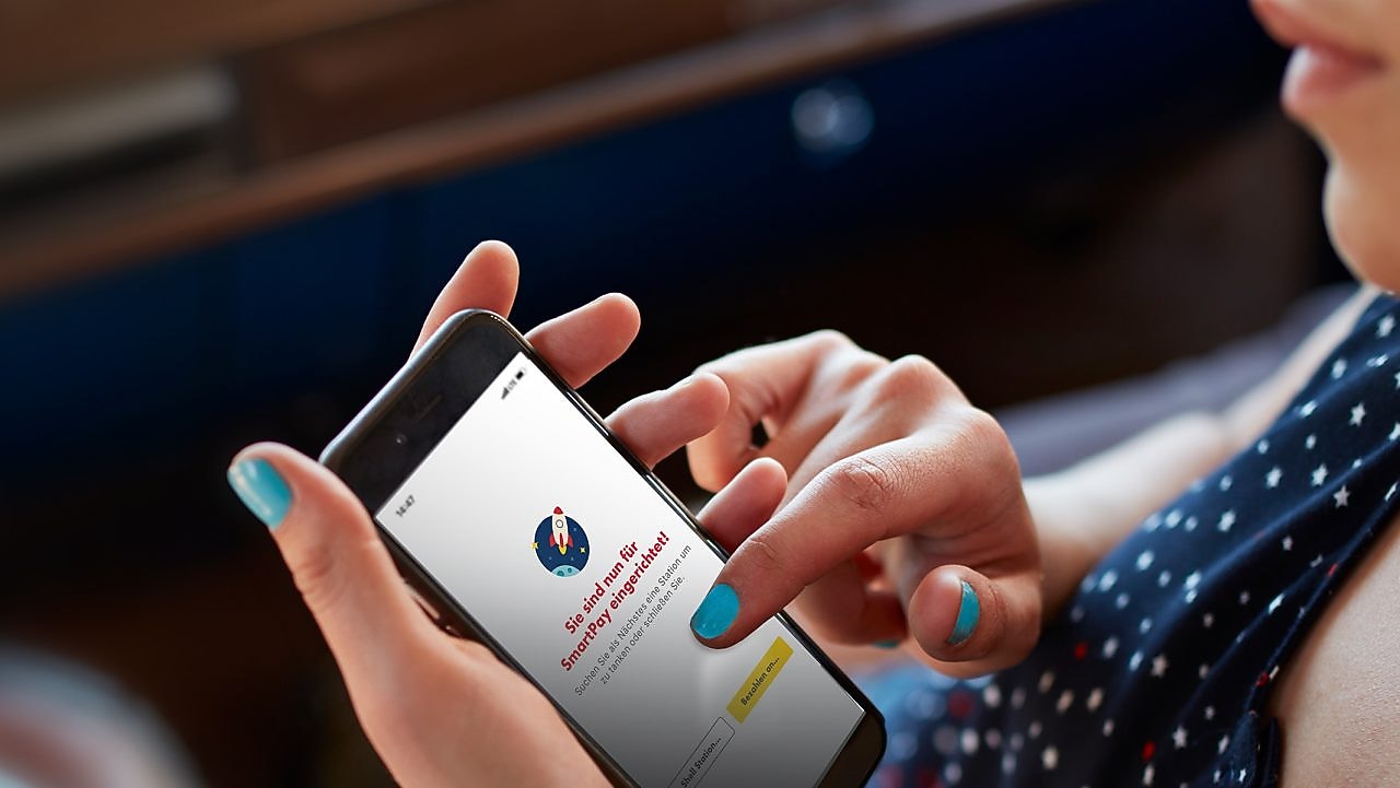 Frau sitzt im Auto und hält in der Hand ein Handy mit geöffneter Shell App und fertig eingerichtetem Shell SmartPay