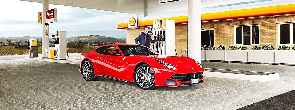 Ein roter Ferrari auf dem Vorplatz einer Shell Station mit einem Mann, der an einer Zapfsäule lehnt