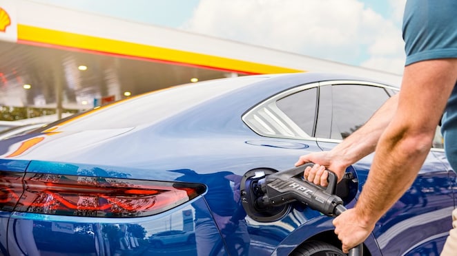 Mann trinkt einen Kaffee, während ihr Elektroauto an einer Shell Recharge Ultra-Schnellladesäule lädt