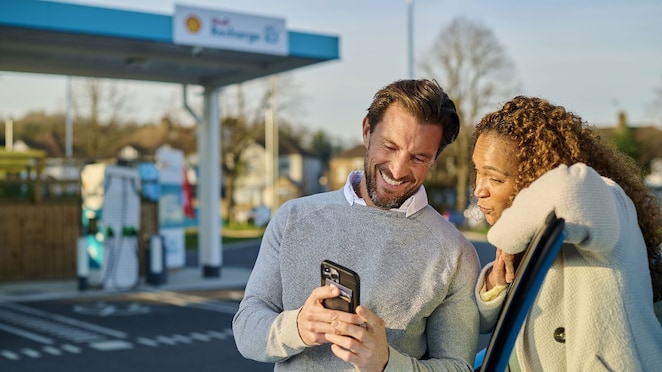 Ein Mann und eine Frau stehen lächelnd vor einer Shell Recharge Ladestation und schauen gemeinsam auf ein Smartphone. Im Hintergrund sind mehrere türkisfarbene Ladesäulen zu sehen.