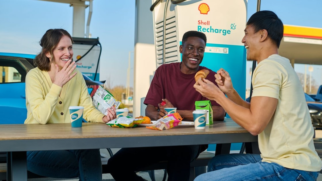 Drei Personen verbringen eine Pause an einer Shell Recharge E-Ladestation. Sie sitzen an einem Tisch mit Getränken und Snacks, während ein blaues Elektrofahrzeug im Hintergrund geladen wird.