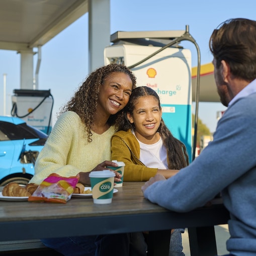 Eine Frau und ein Kind sitzen lächelnd an einem Tisch im Freien an einer Shell Recharge Ladestation. Ein Mann sitzt ihnen gegenüber. Im Hintergrund ist ein Elektrofahrzeug an einer Ladesäule zu sehen.