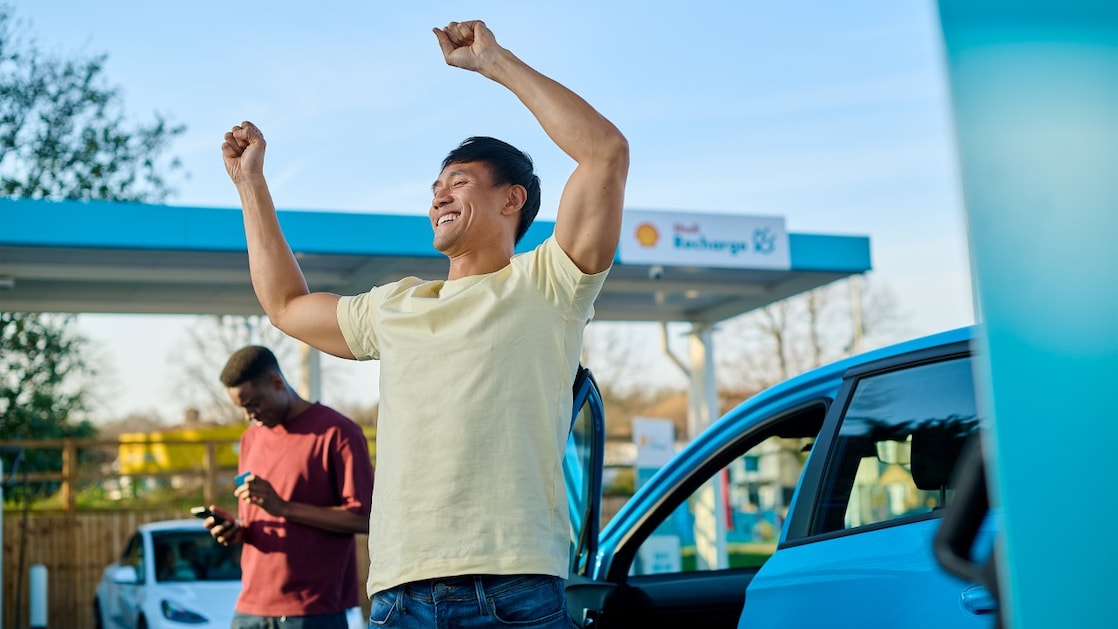 Eine Person steht neben einem Elektroauto mit geöffneter Fahrzeugtür und hebt die Arme. Im Hintergrund befindet sich eine Shell Recharge E-Ladestation, während eine weitere Person neben dem Fahrzeug auf ein Smartphone blickt.