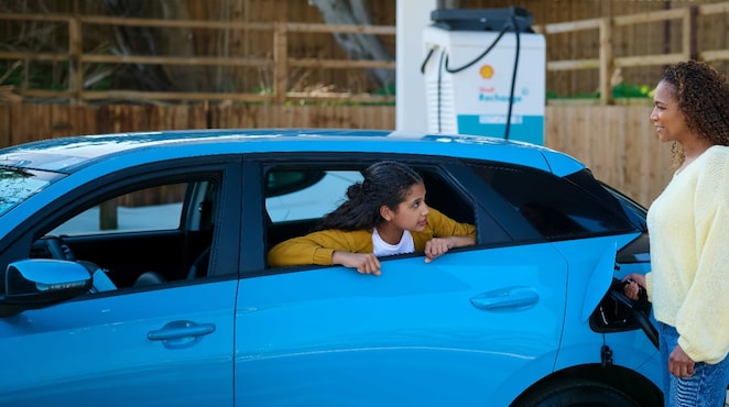 Frau lädt ein blaues Elektroauto an einer Shell Recharge Ladesäule, während ein Kind aus dem geöffneten Autofenster schaut