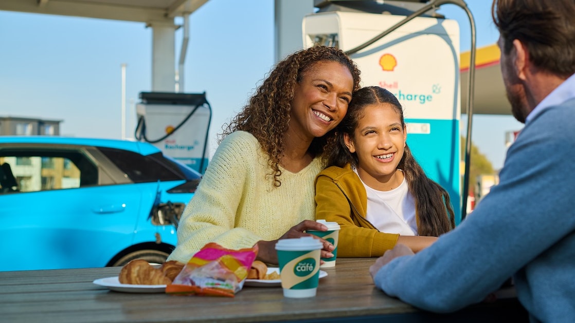 Personen machen eine Pause an einer Shell Recharge E-Ladestation. Im Hintergrund wird ein blaues Elektrofahrzeug geladen.