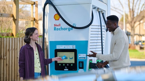 Eine Frau und ein Mann stehen vor einer Shell Recharge Ladesäule. Die Frau bedient das Display der Ladesäule. Im Hintergrund ist ein Fahrzeug zu sehen.