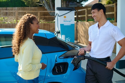 Zwei Personen stehen neben einem blauen Elektrofahrzeug an einer Shell Recharge E-Ladestation. Eine Person hält ein Ladekabel in der Hand.​