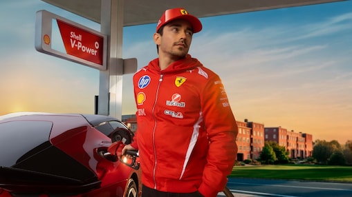 Formel-1-Fahrer Charles LeClerc&nbsp;in rotem Rennanzug betankt einen roten Sportwagen an einer Zapfsäule mit Shell V-Power Beschilderung bei warmem Abendlicht.