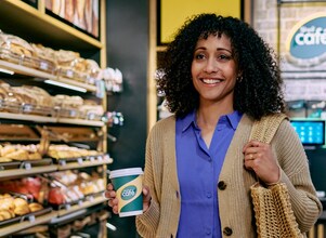 Frau steht lächelnd mit einem Kaffee von Shell Café in einer Shell Station mit Shell Café Backshop